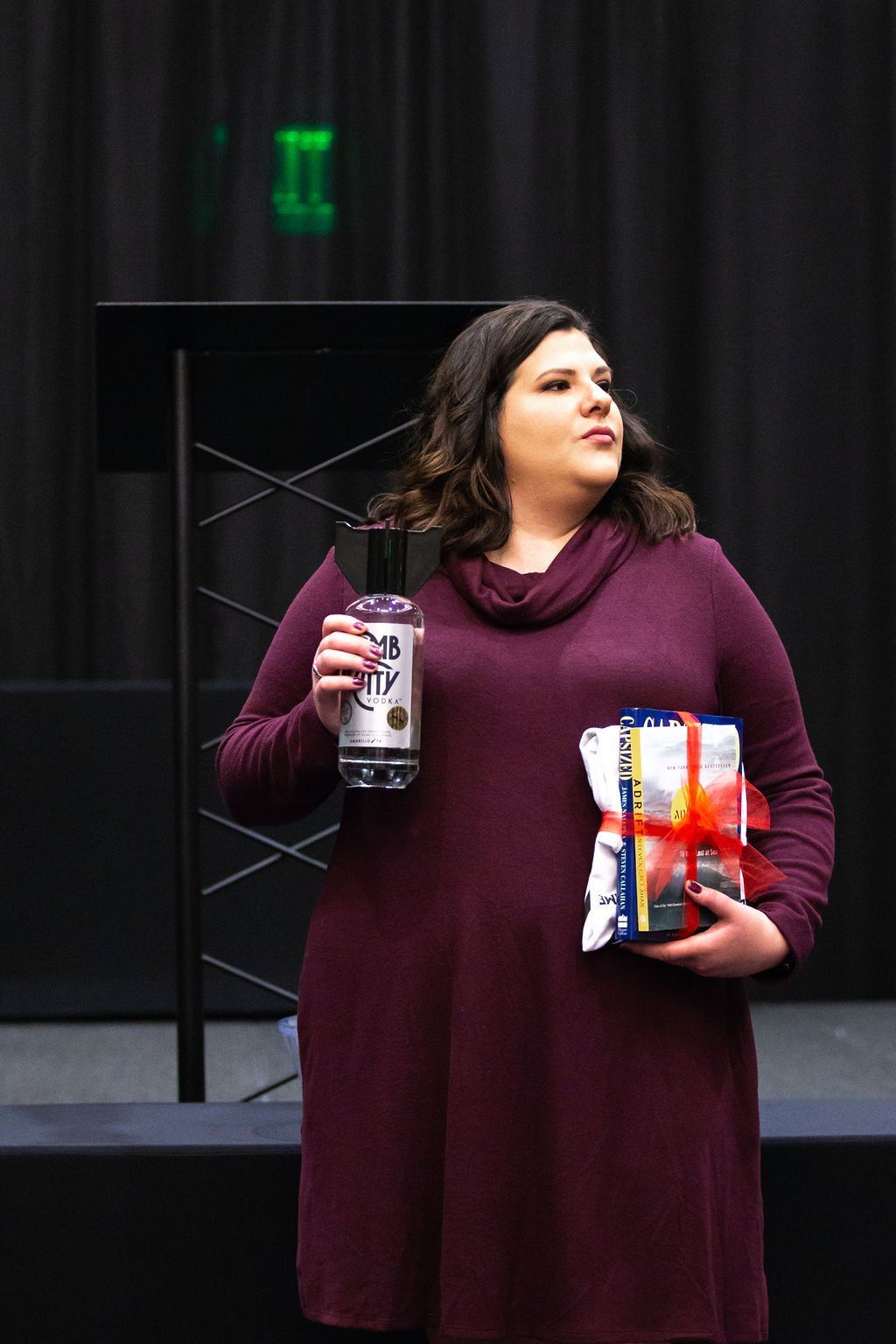 A person in a maroon sweater holding a bottle and a gift-wrapped item against a dark backdrop.