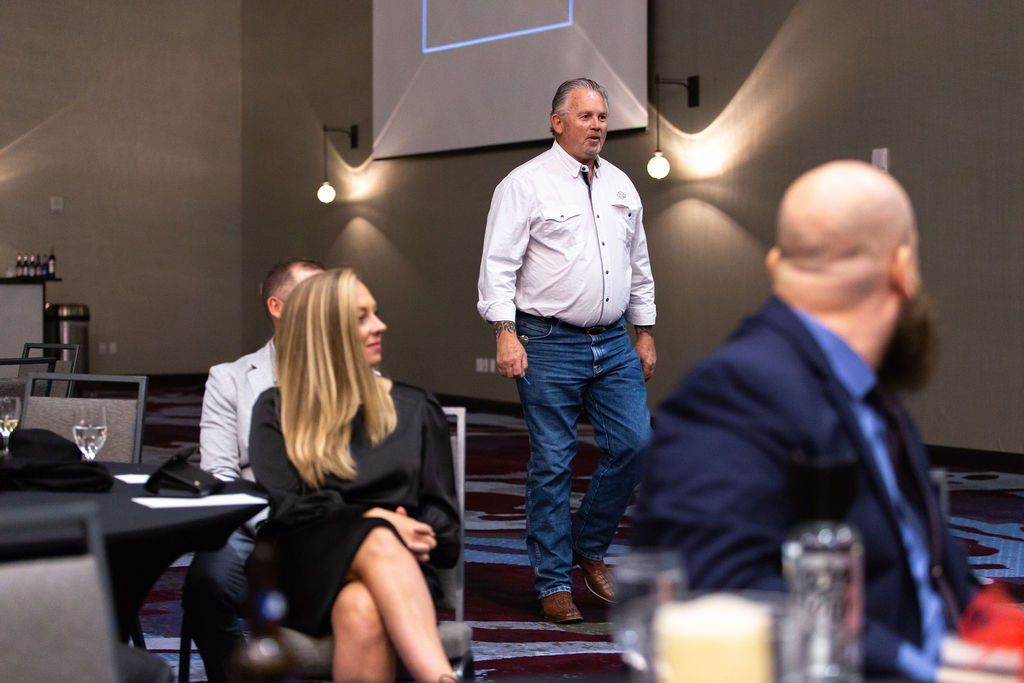 A speaker in a light shirt and jeans stands at the front of a room, addressing an audience sitting at tables.
