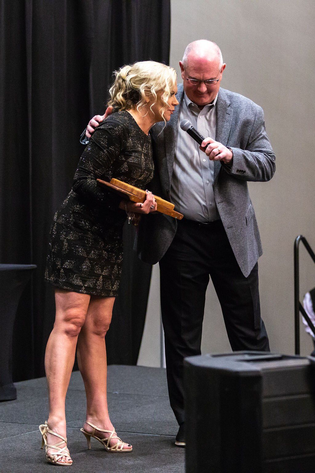 A person in a black patterned dress accepts an award from someone in a grey suit holding a microphone on a stage.