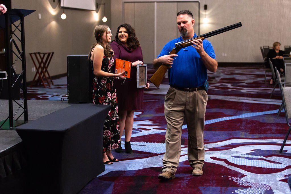 A man in a blue polo holding a rifle, accompanied by two individuals on stage in a carpeted event hall.
