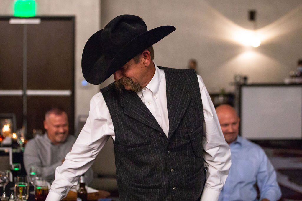 A man in a black cowboy hat and striped vest standing in a room with two people seated in the background.