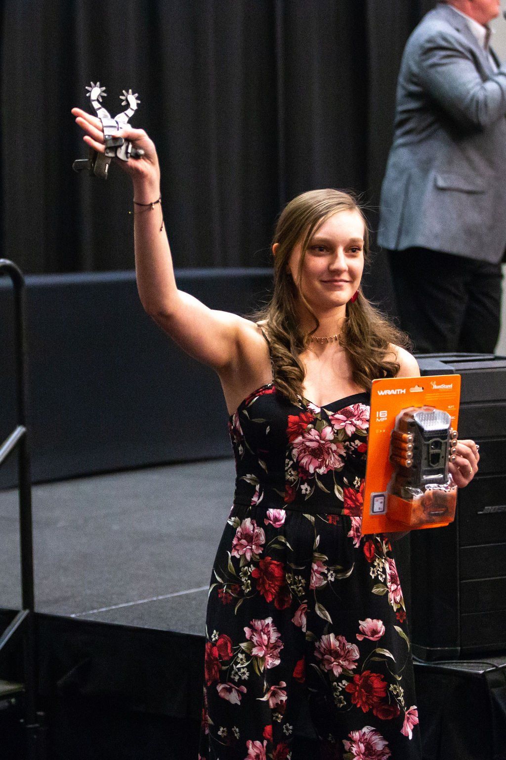 A person wearing a floral dress smiles while holding a pair of spurs aloft and a boxed trail camera on a stage.