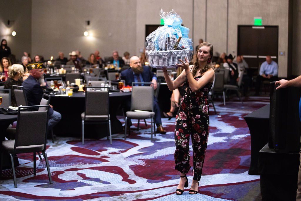 A person in a floral jumpsuit carries a large gift basket wrapped in blue cellophane through a banquet hall.