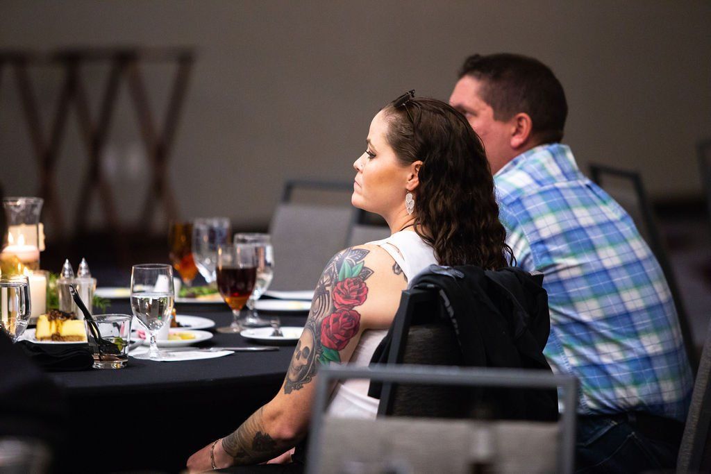 A person with a large floral arm tattoo and a companion in a plaid shirt sit at a dinner table in a banquet hall.