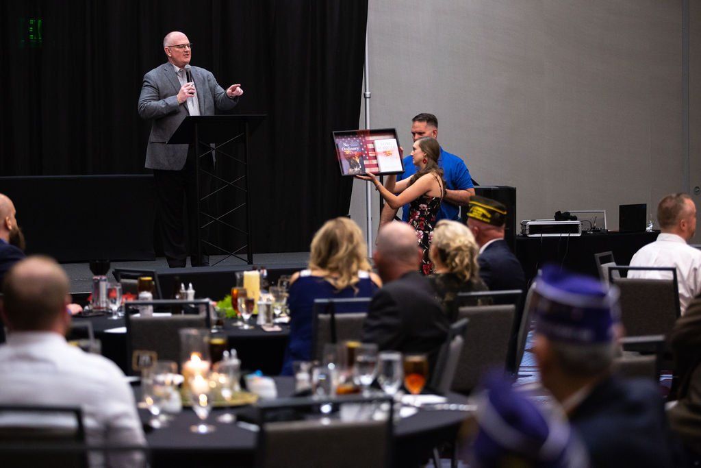 A speaker stands at a podium in a banquet hall while a presenter holds a framed certificate for the audience to view.