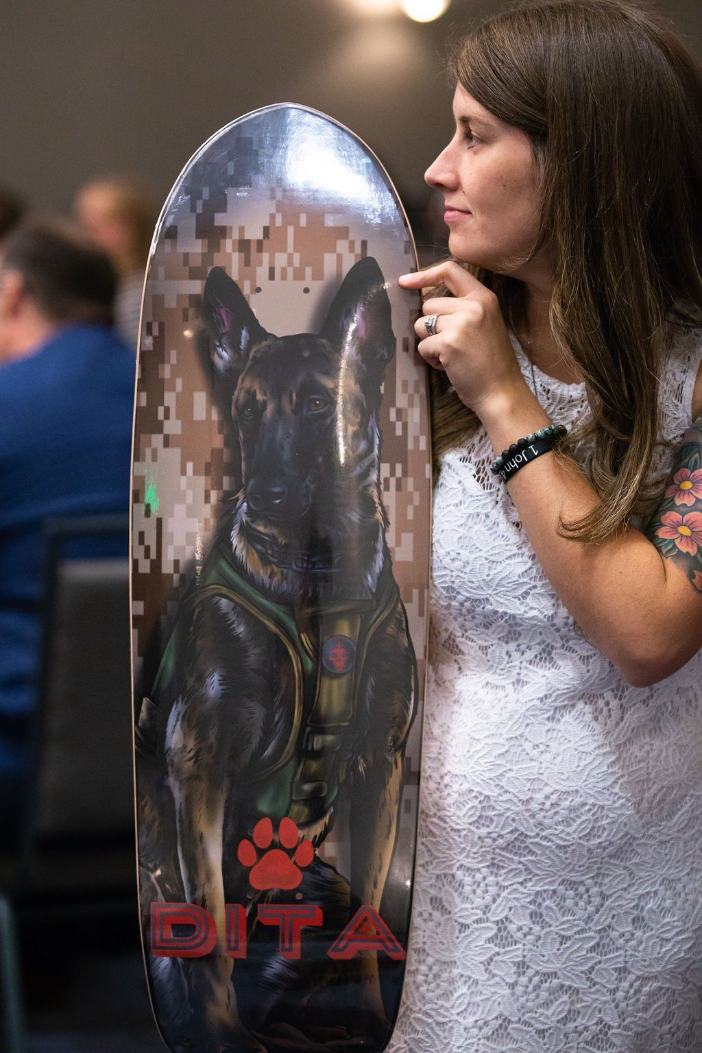 A person in a white dress holds a vertical skateboard deck featuring a portrait of a military dog with a paw print logo.