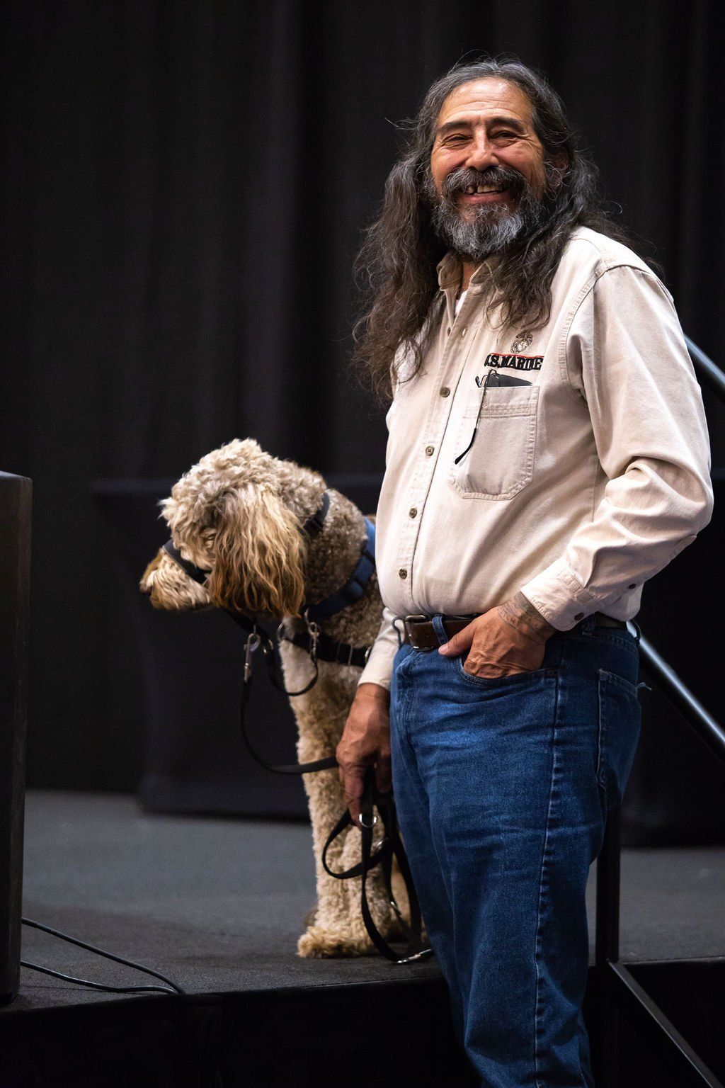 A smiling person with long dark hair stands on a stage next to a curly-coated dog wearing a harness and leash.
