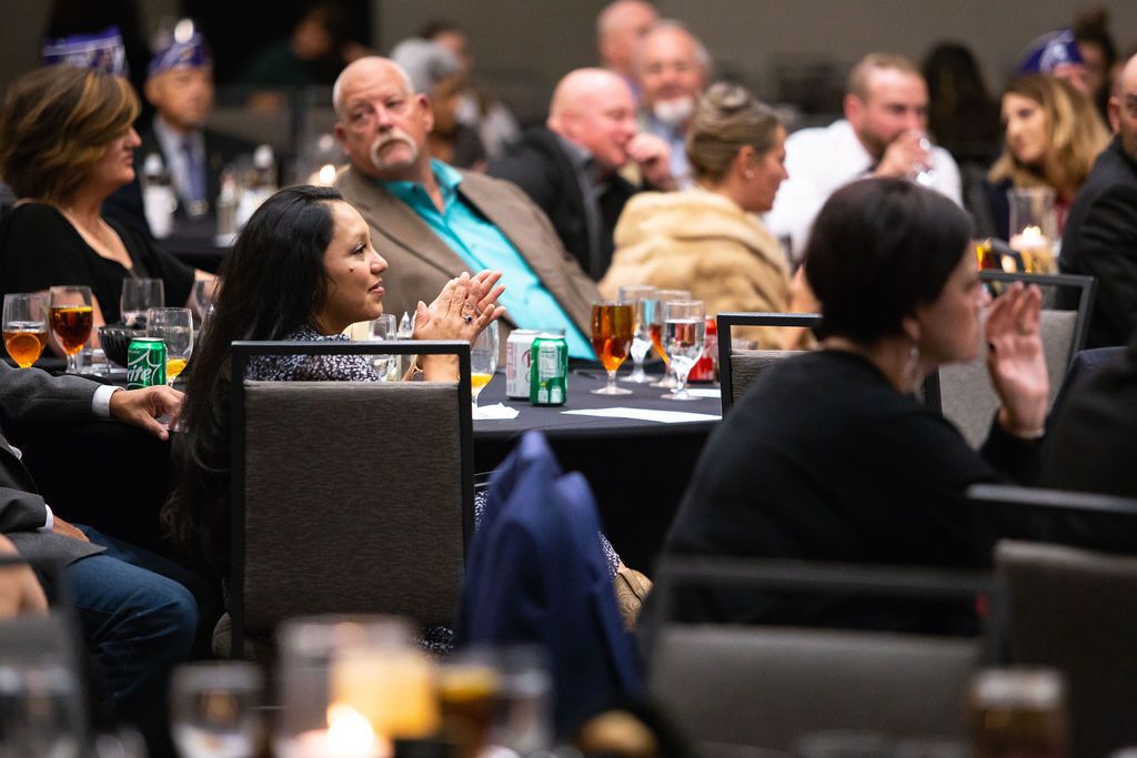 People sit at tables in a dimly lit banquet hall, with some attendees clapping during a formal event.