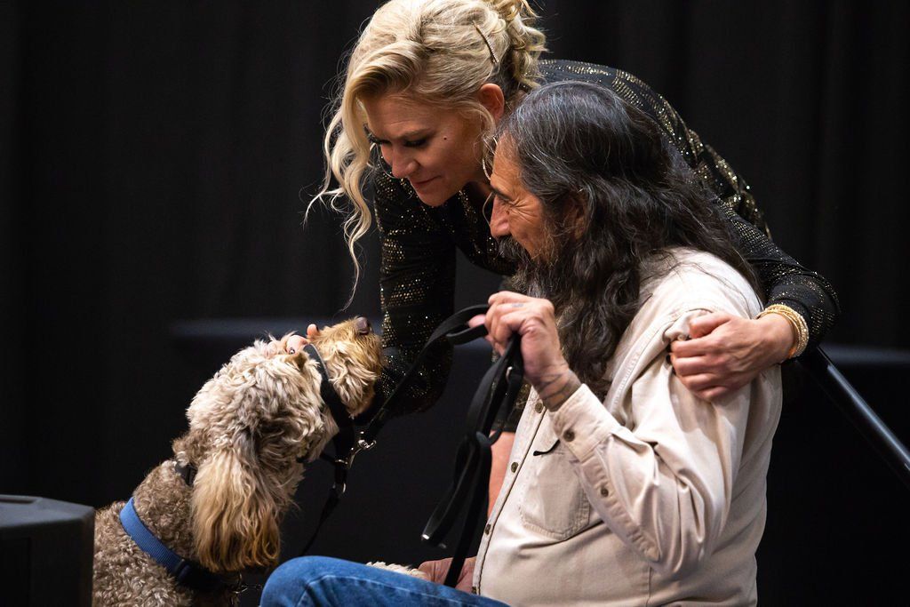 A woman with blonde hair places a hand on a man's shoulder while he interacts with a dog wearing a blue collar.