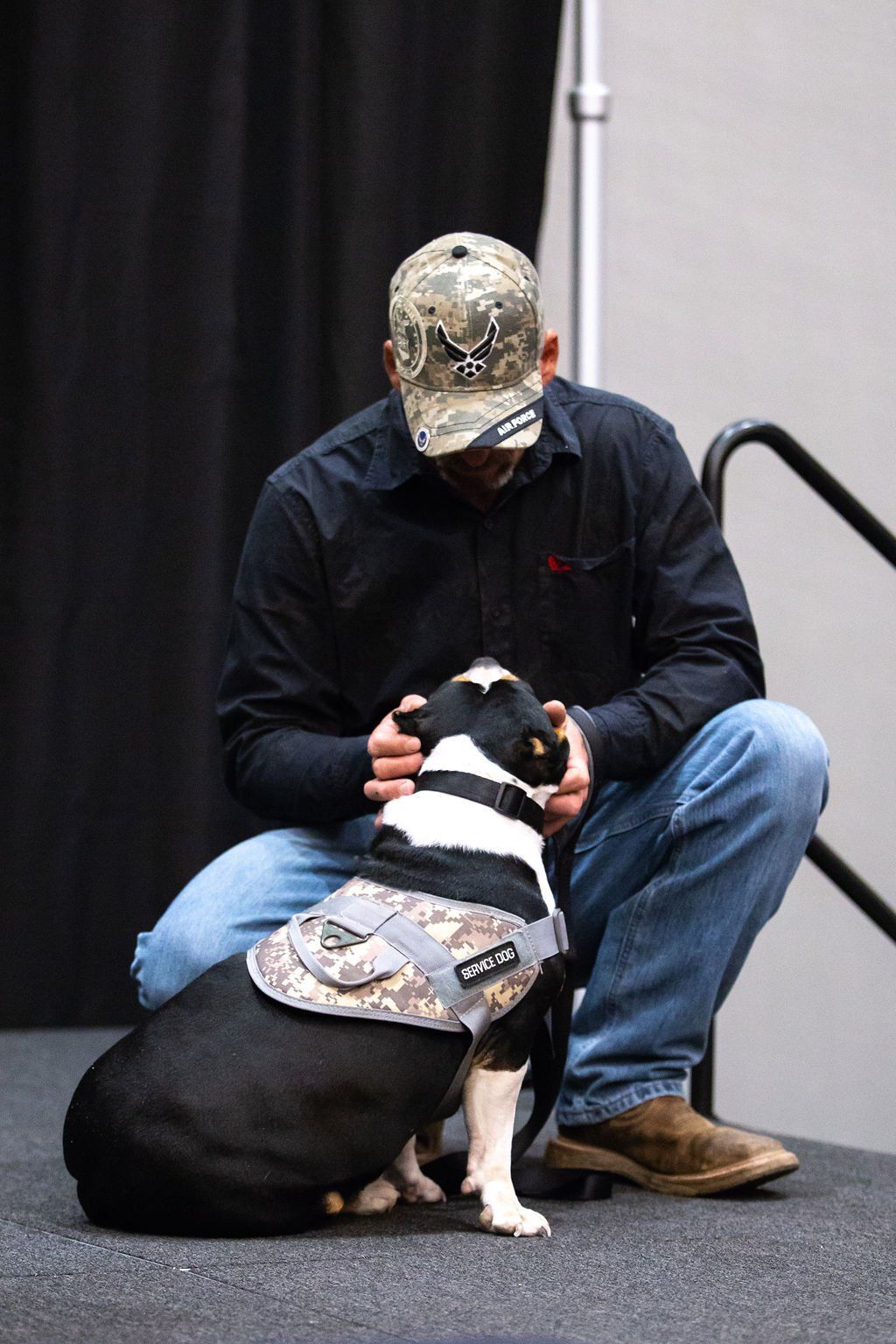 A person in a camouflage cap and dark shirt kneels to pet a black and white dog wearing a matching camouflage harness.