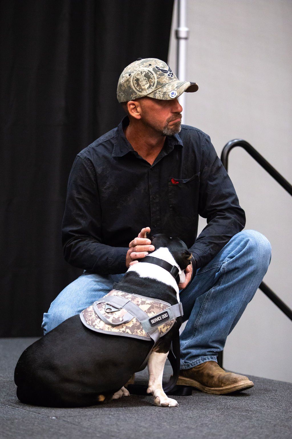 A person in a camouflage cap and dark shirt kneels on a stage, gently petting a dog wearing a matching camo vest.