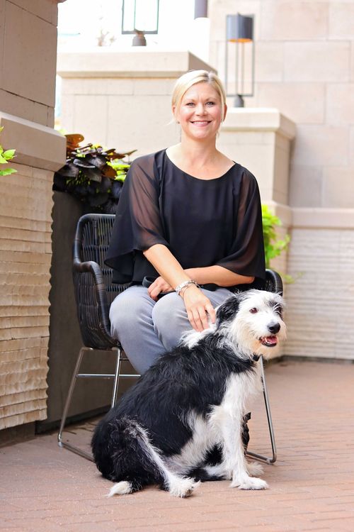 A blonde woman in a black shirt and patterned pants sits on a chair next to a black-and-white dog on a stone patio.