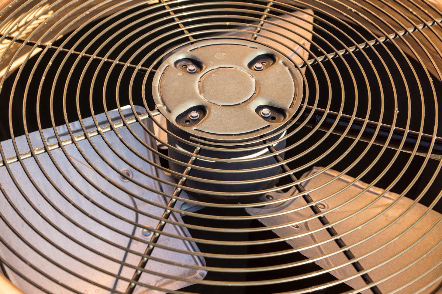 Metal fan blades behind a circular wire grill. The fan is likely part of an air conditioning unit.