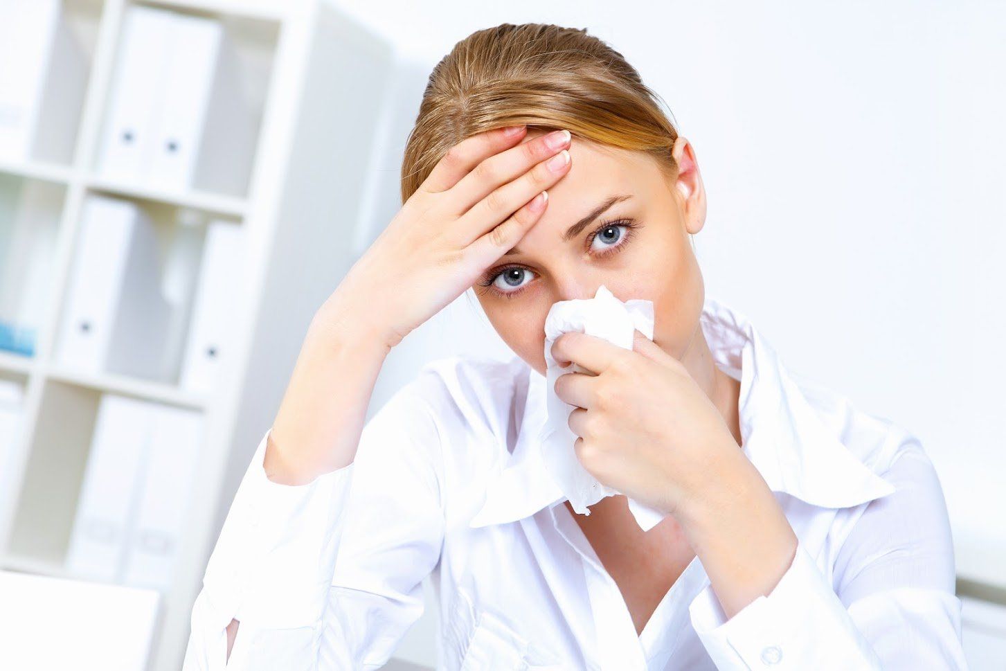 Woman in a white shirt with a tissue to her nose, touching her forehead, indoors.
