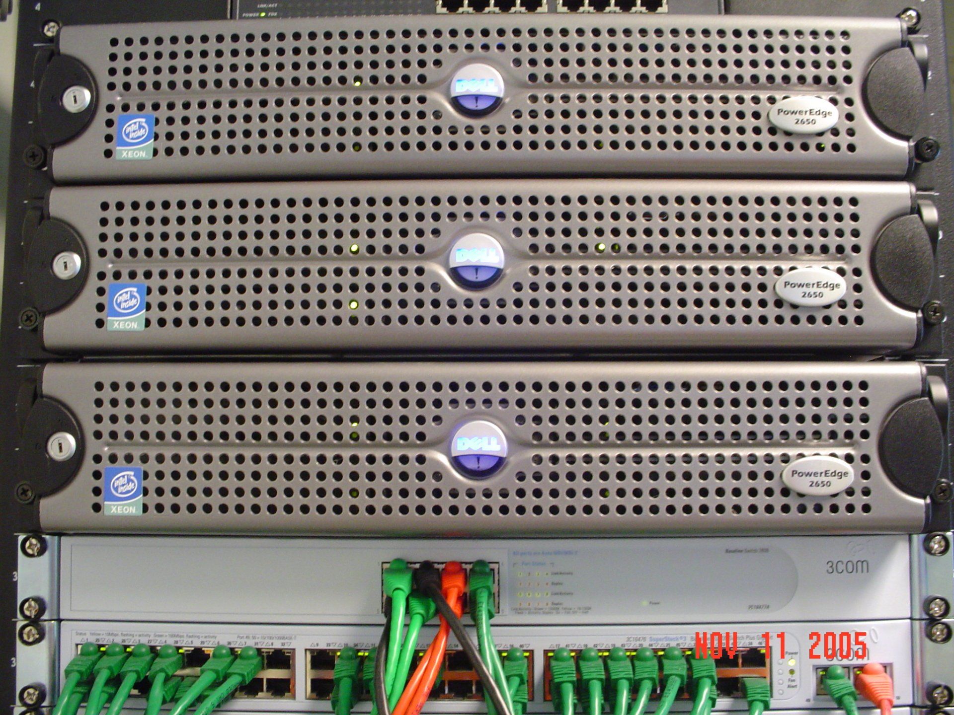 Three gray server units stacked above a network switch, connected by green and orange cables.
