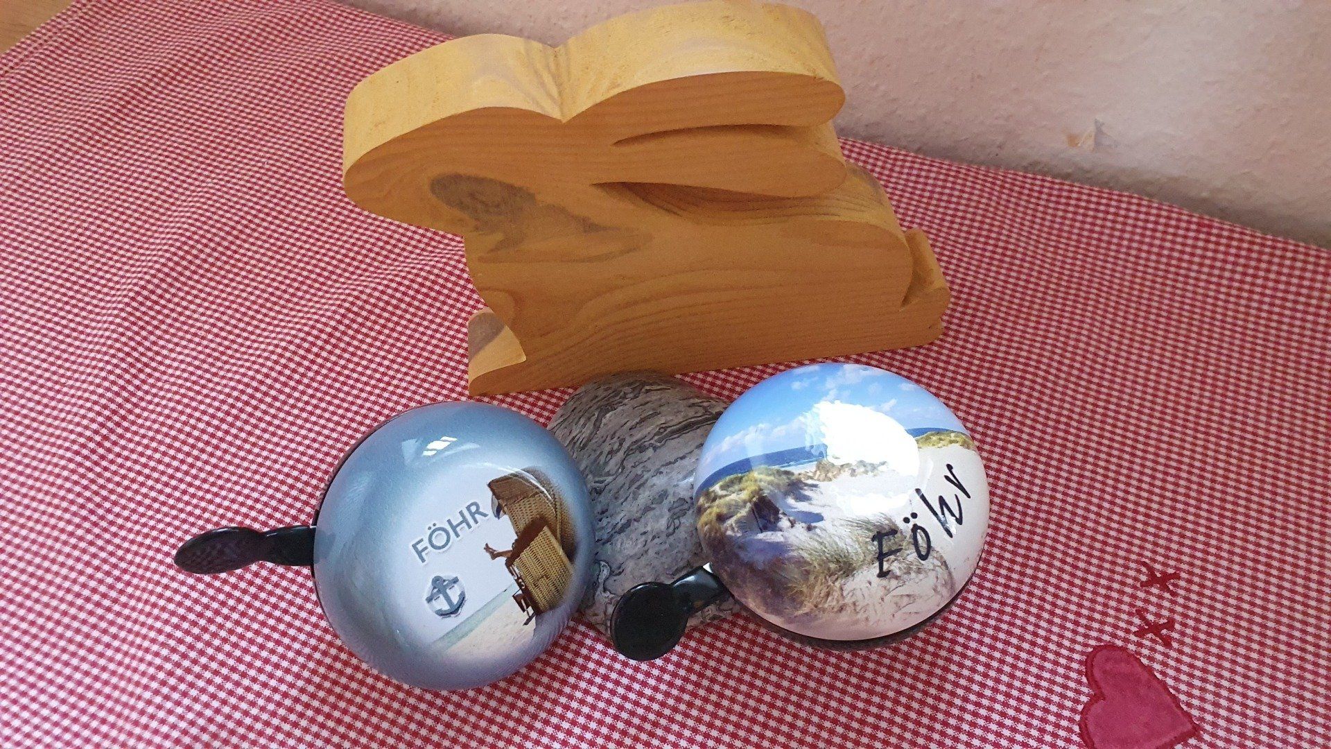 Two blue and white bells with pictures and text, and a wooden heart-shaped carving, sit on a red and white patterned surface.