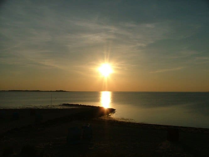 Sun setting over calm water, with a beach in the foreground and a hazy sky.