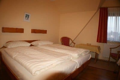 Bedroom with a double bed, bedside table, two chairs, and a window with red curtains. Beige walls.