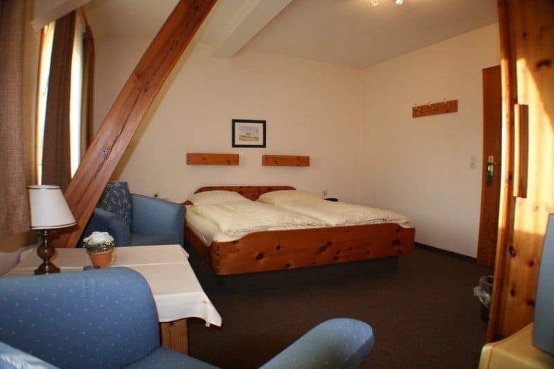 Bedroom with a double bed, blue armchairs, wooden beams, and a table. Brown curtains and a lamp are visible.