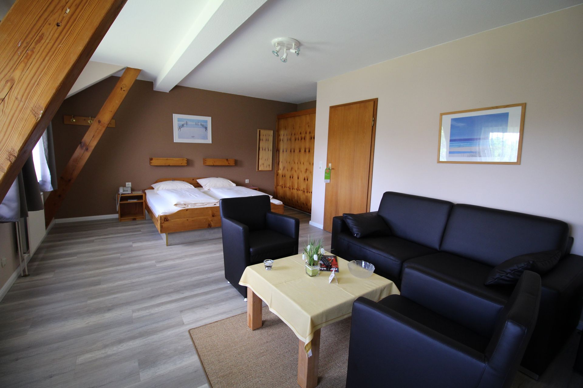 Bedroom with bed, sofa, chairs, and wooden beams. Brown and white walls, light wood floor, and a small table.