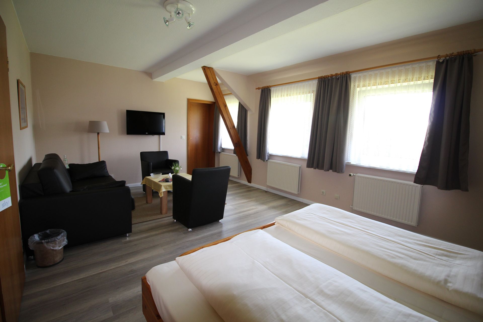 A hotel room with two beds, a sofa, chairs, and a TV. The room has windows with curtains and a wooden beam.