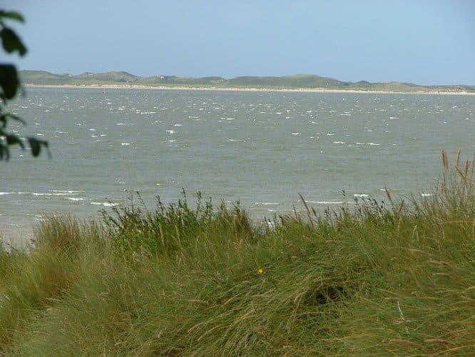 Grassy dunes in the foreground frame a wide, choppy sea with a distant shoreline and overcast sky.