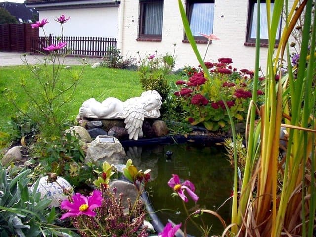 A garden pond with a white angel statue, colorful flowers, and reeds in front of a house.
