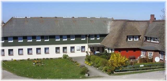A building with a gray roof and windows sits next to a red building with a thatched roof and green grass in the foreground.