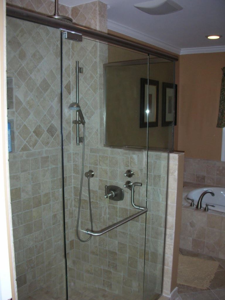 Glass-enclosed shower with beige tile walls and fixtures.  A chrome handrail, showerhead, and controls are visible.