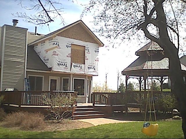House exterior under construction, Tyvek wrap, with a deck and gazebo in the backyard on a sunny day.