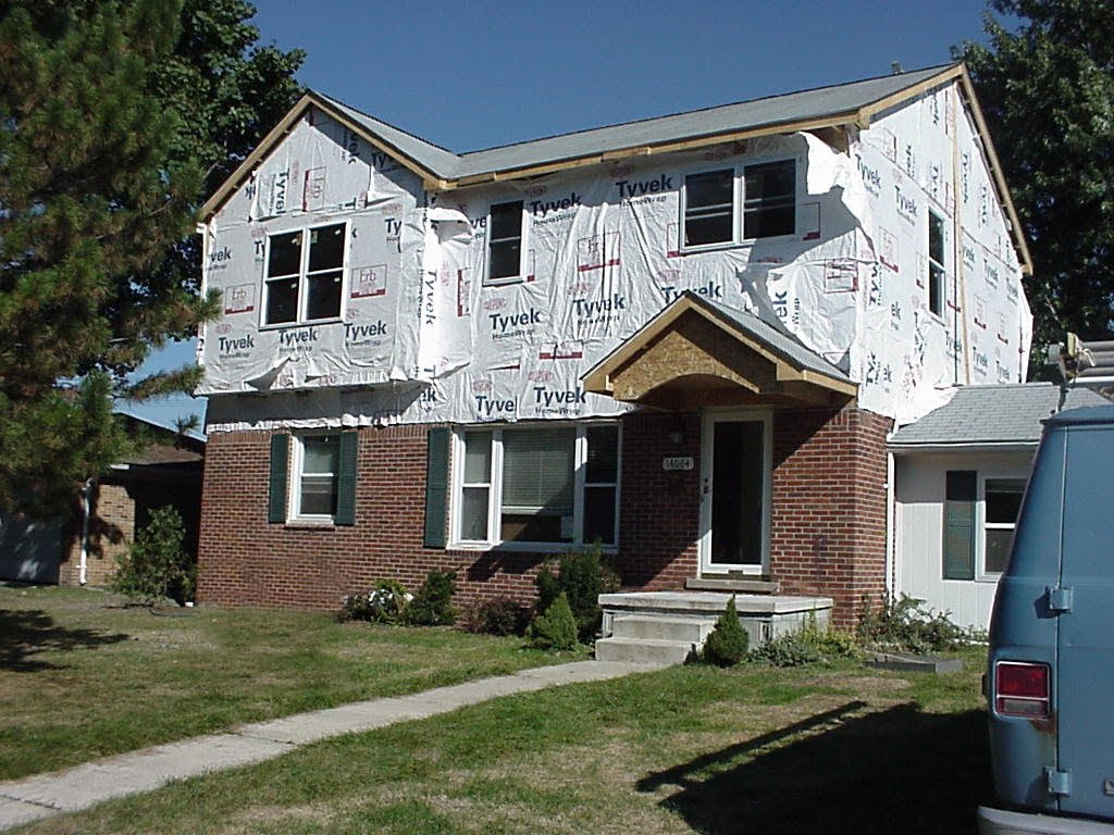 Two-story house undergoing renovation; sheathing on upper level, brick on lower, blue van on right.