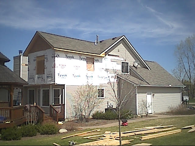 Two-story house undergoing siding replacement; Tyvek wrap visible. Lumber on the ground, sunny day.
