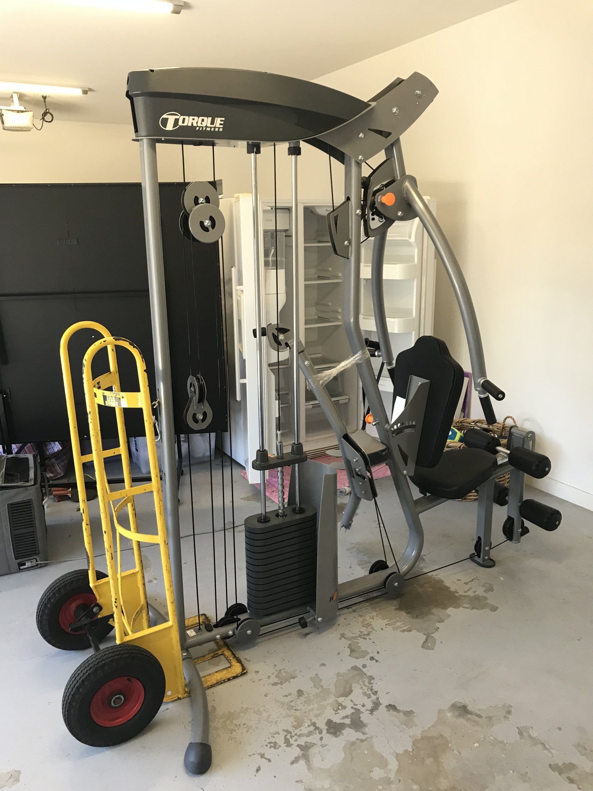 A gym machine is sitting in a garage next to a yellow dolly.