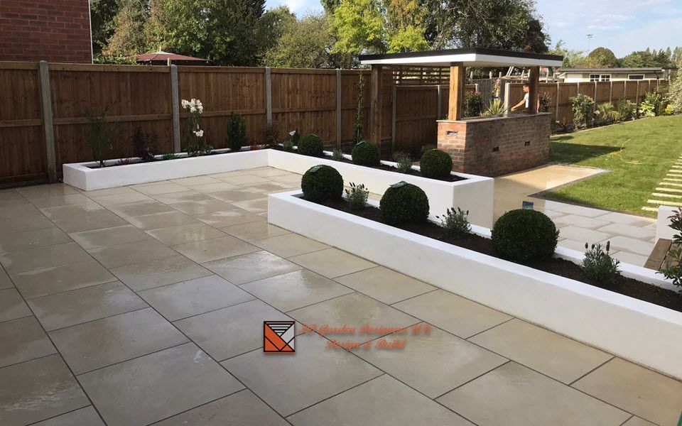 Paved patio with white planters and trimmed shrubs, wooden fence, gazebo, and green lawn.