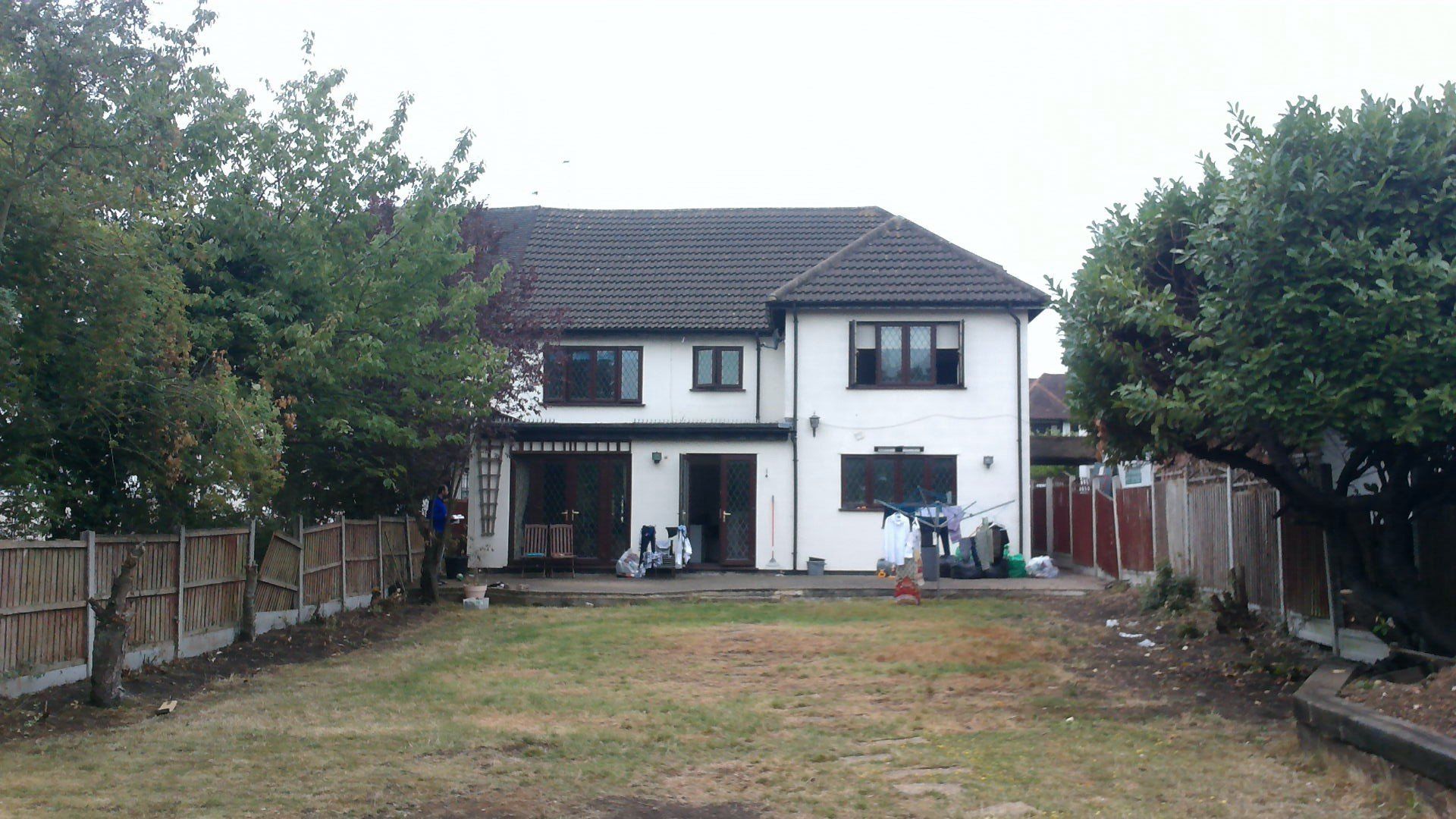 Two-story white house with thatched roof, brown wooden fence surrounds backyard with overgrown grass.