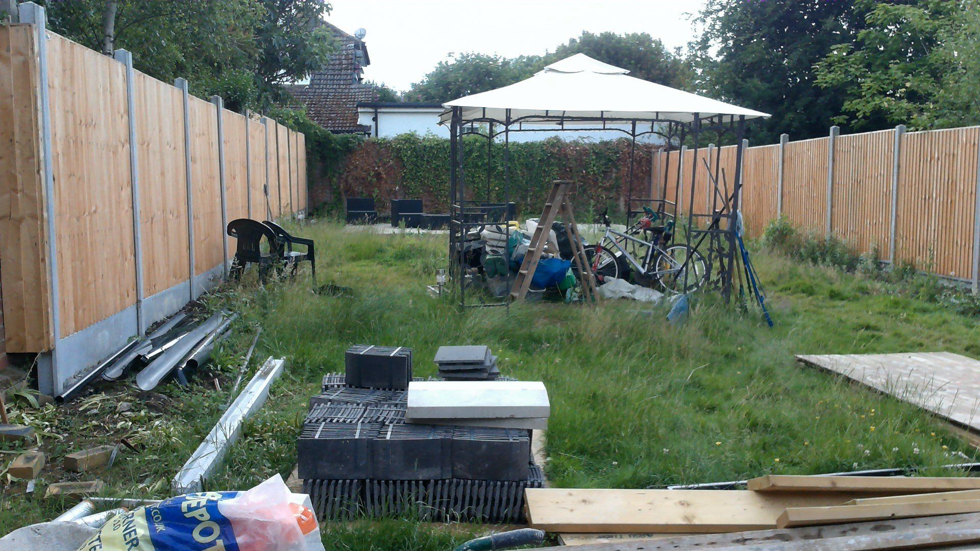 Backyard with a wooden fence, gazebo, and overgrown grass with construction materials.