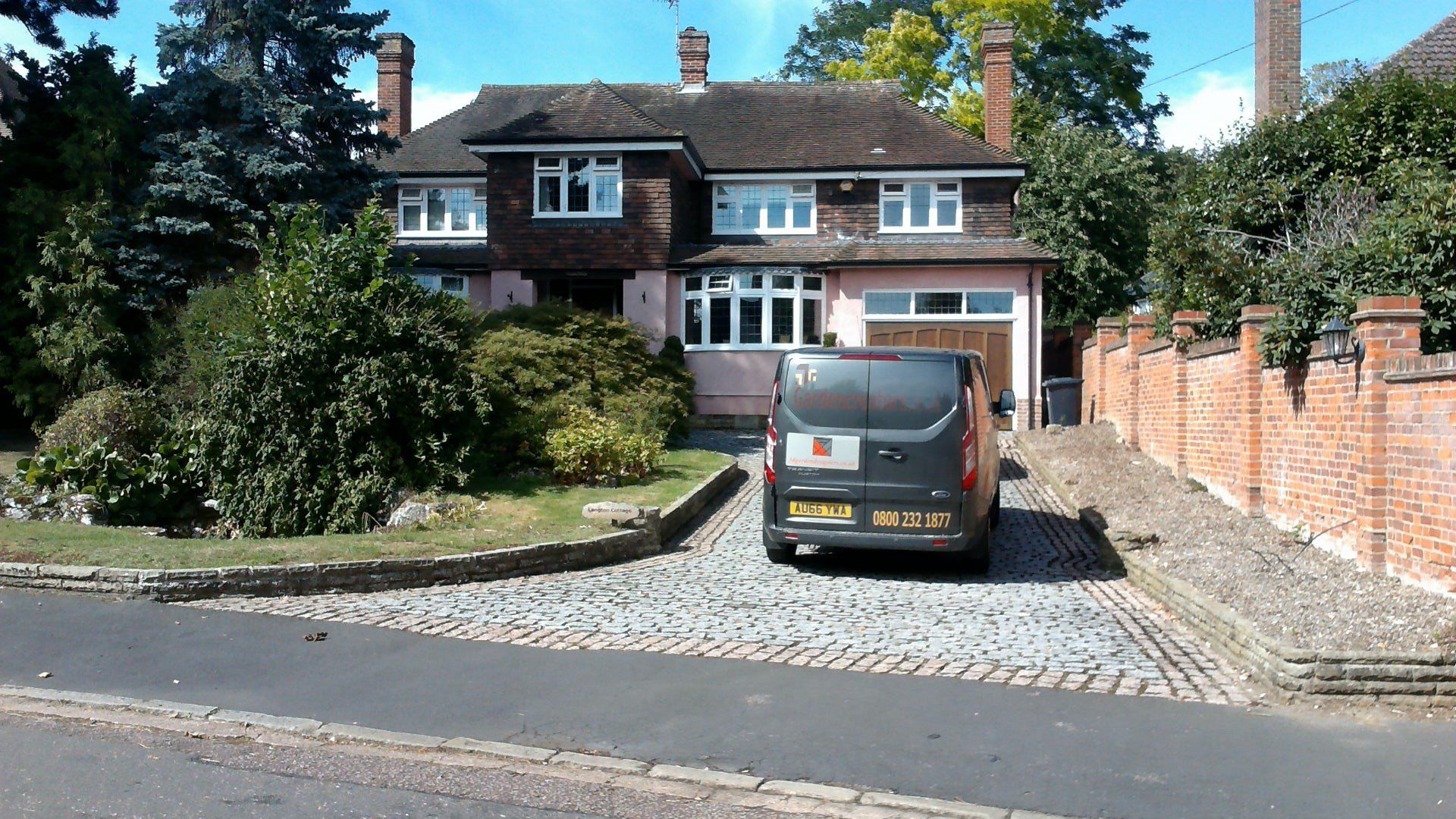 before House with pink walls, brown roof, and van parked in cobblestone driveway. Brick wall on right.
