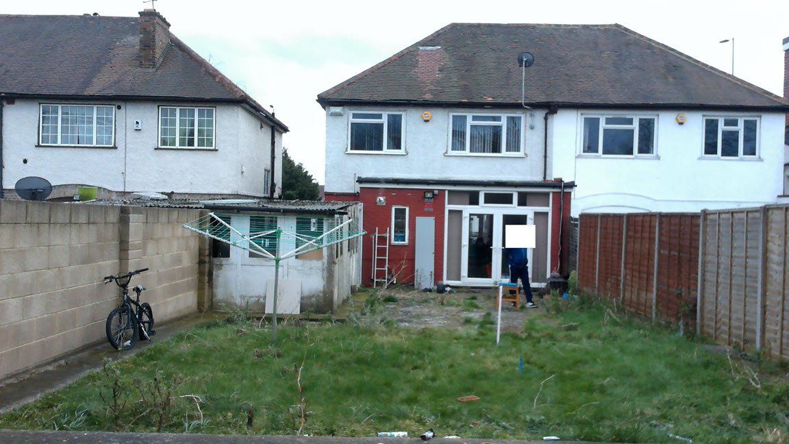 Backyard view of a house with overgrown grass, a bicycle, and a fence. A person is visible near the back door.