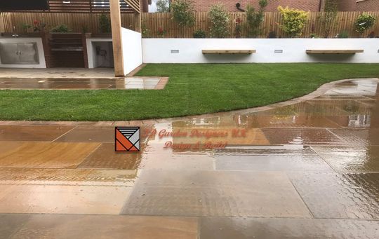 Wet stone patio and green lawn with built-in benches, white wall, and a wooden fence; an orange and black sign sits on the patio.