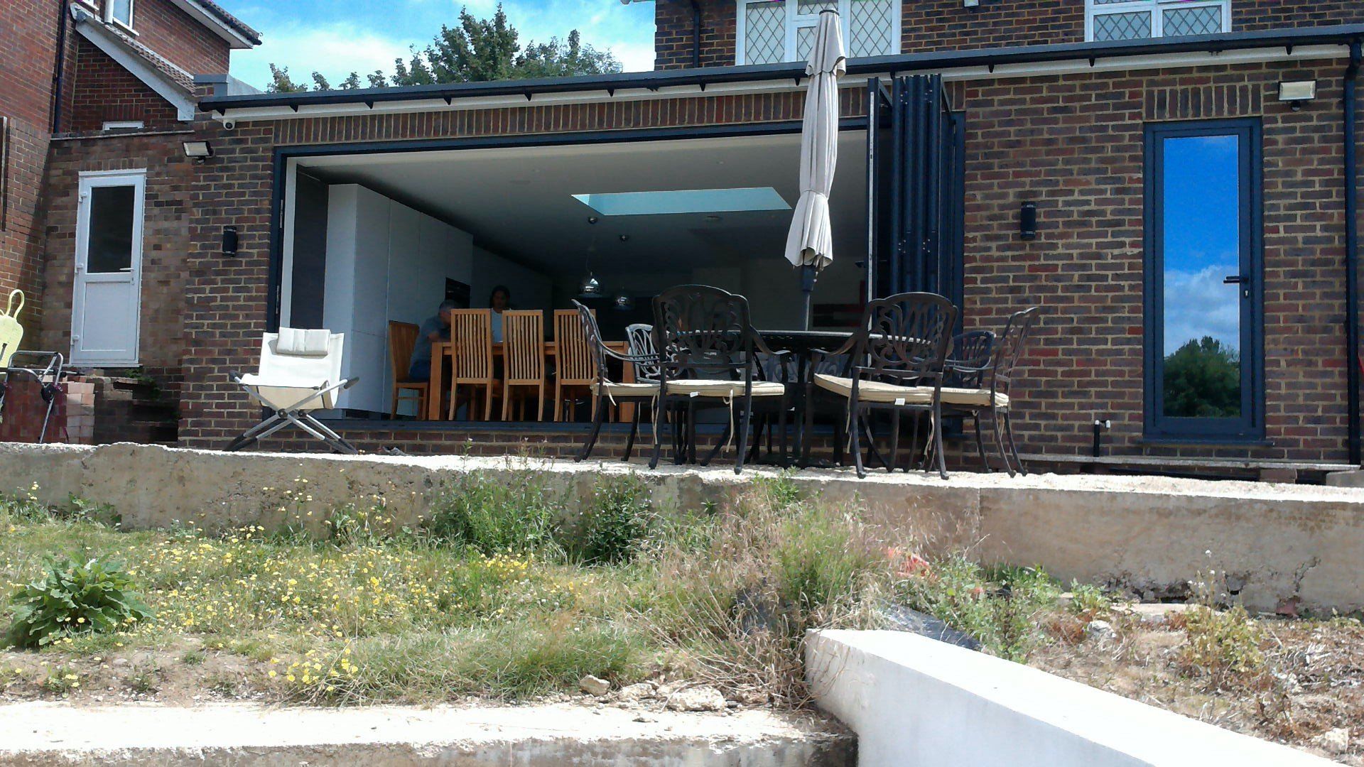 Brick house exterior with open sliding doors, revealing an interior dining area, patio furniture, and overgrown foreground.