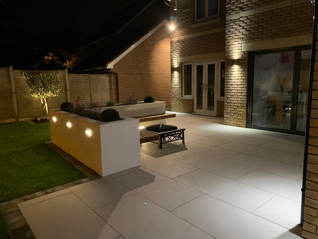 Lit patio at night with modern landscaping. Brick house, square paving stones, and built-in planters with warm lighting.