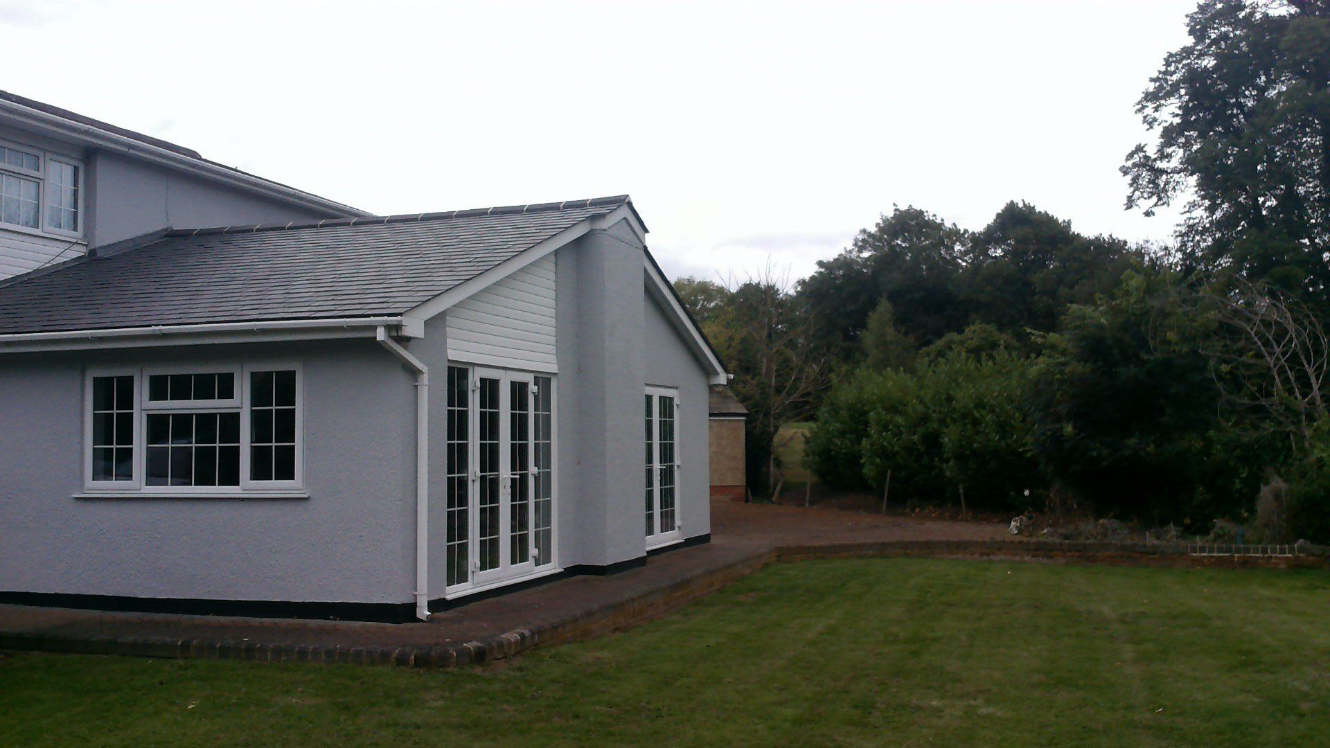 Light gray house extension with white trim, windows, and glass doors, set against a green lawn and trees.