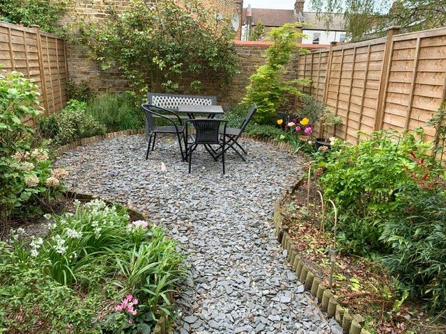 Gravel patio with a table and chairs surrounded by flower beds, wooden fence, and greenery.