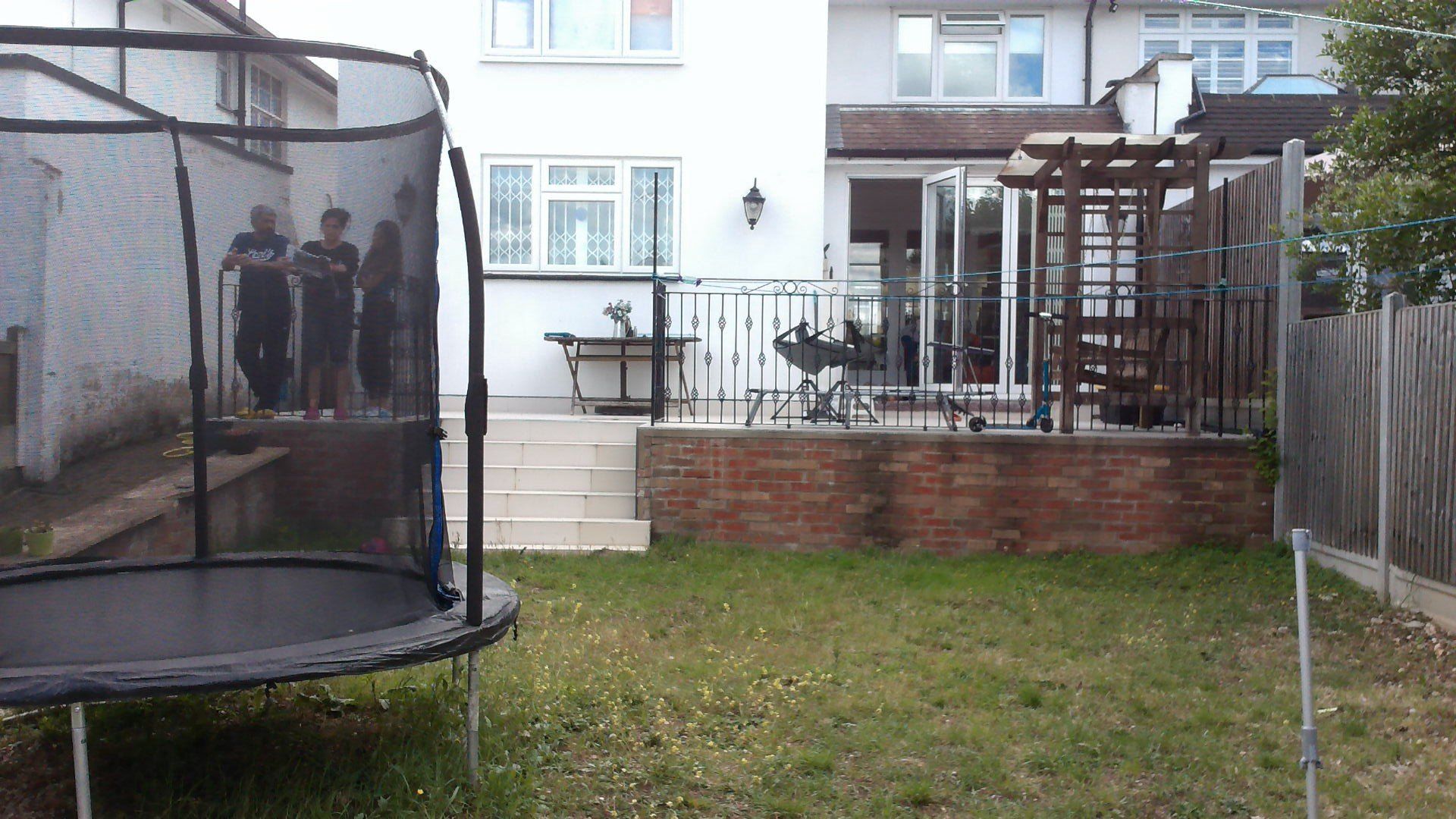 Trampoline in backyard with three figures near it, brick patio, and white house.