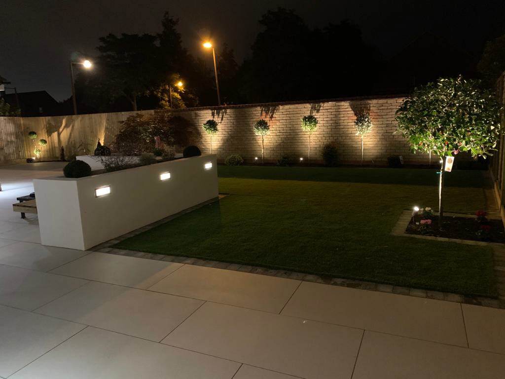 A well-lit, modern backyard at night with a manicured lawn, stone patio, and illuminated trees.