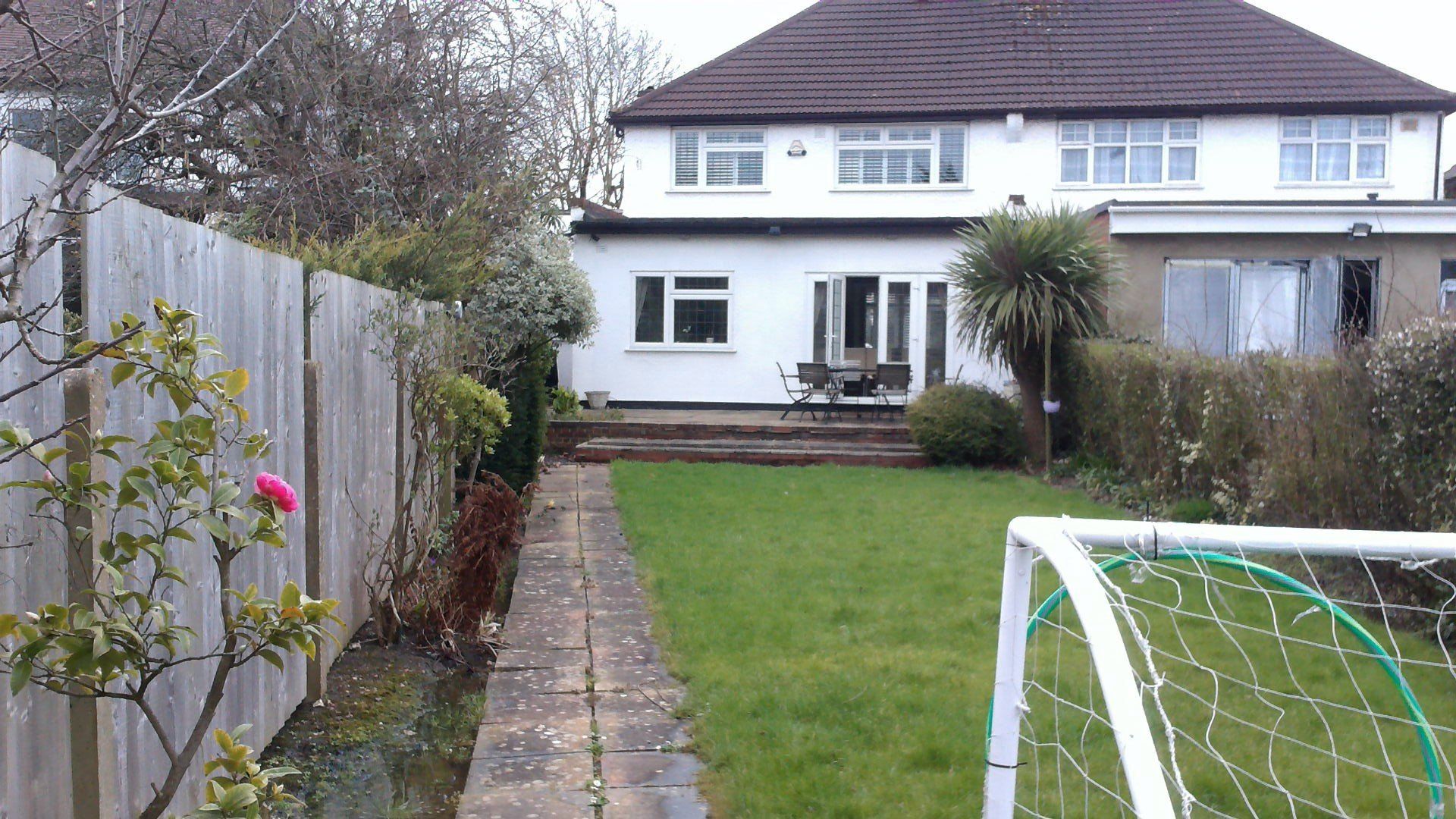 Backyard with green lawn, paved path, white house, wooden fence, and a soccer goal.