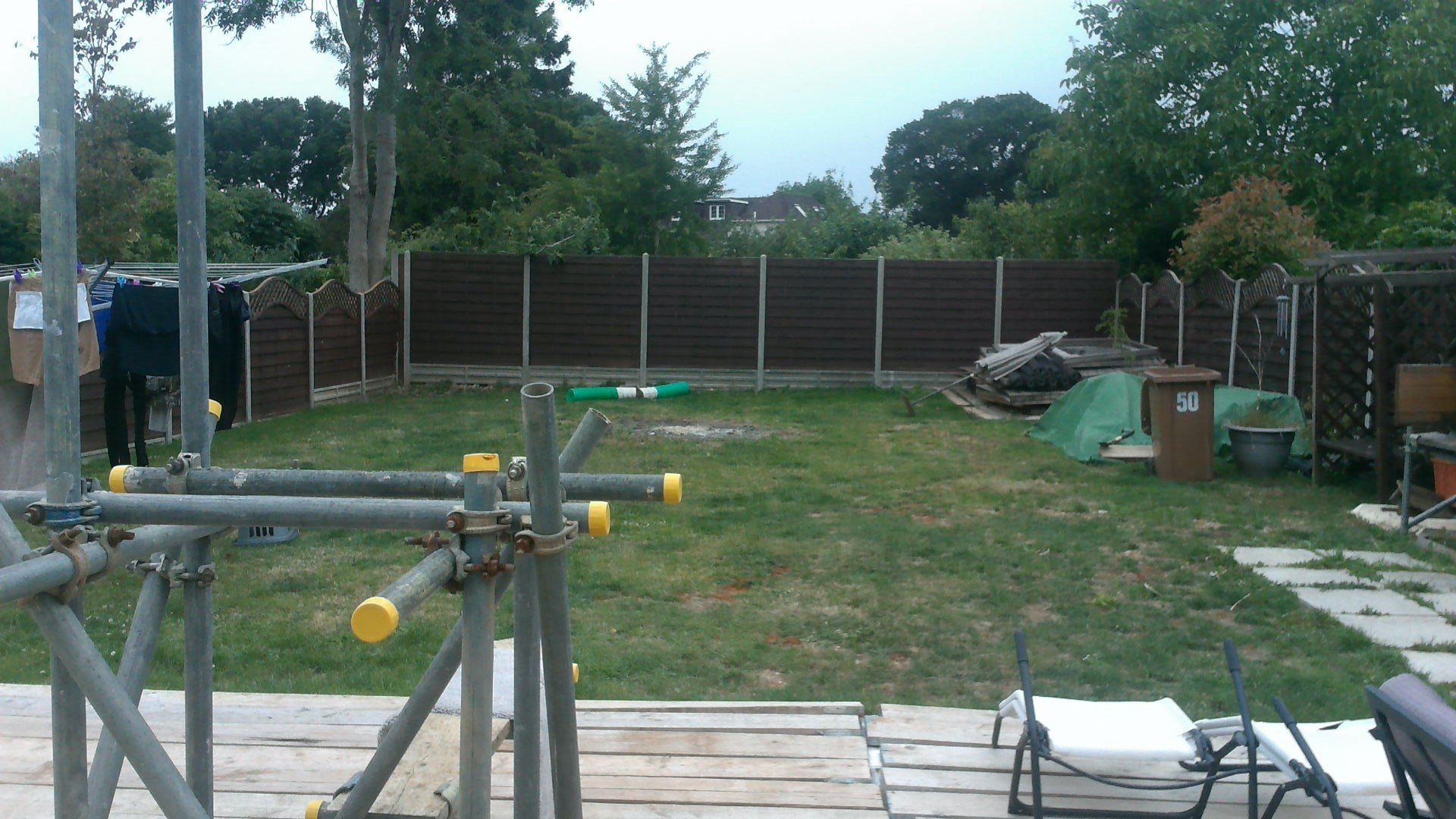 Backyard with a brown fence, scaffolding in foreground, and grass.