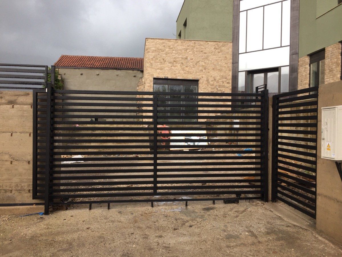 Black horizontal slat metal gate in front of buildings, on a dirt surface.