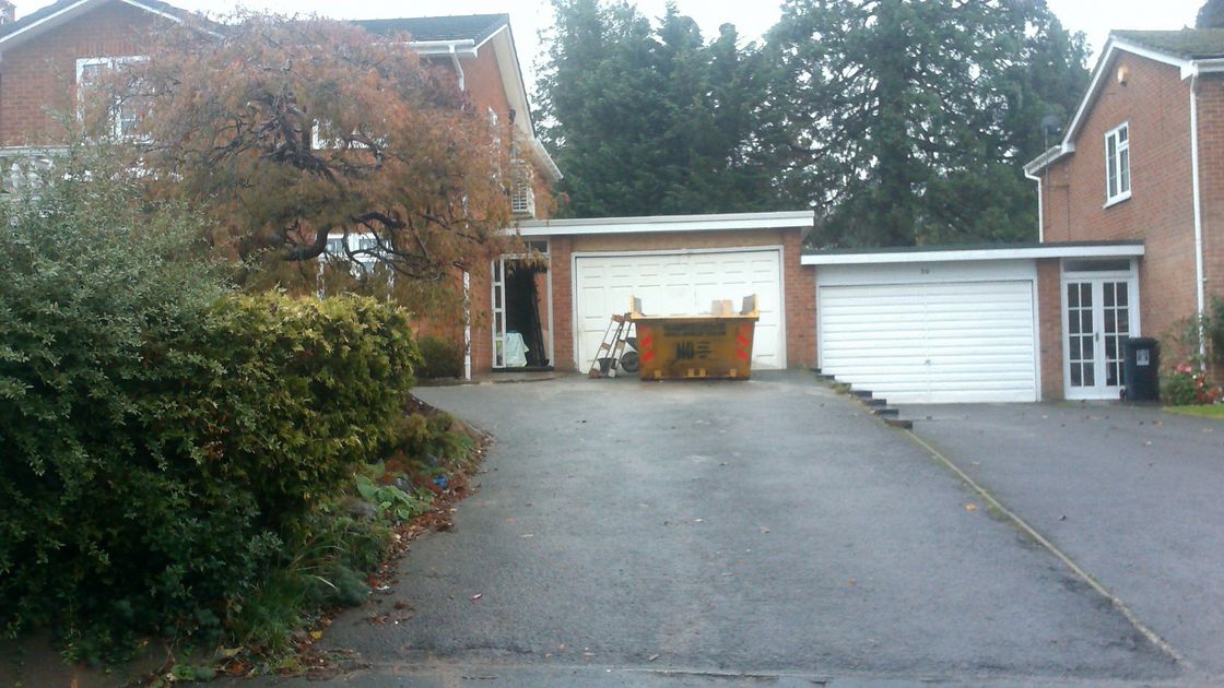 Driveway leading to garages, a two-story house with overgrown ivy, and a skip.