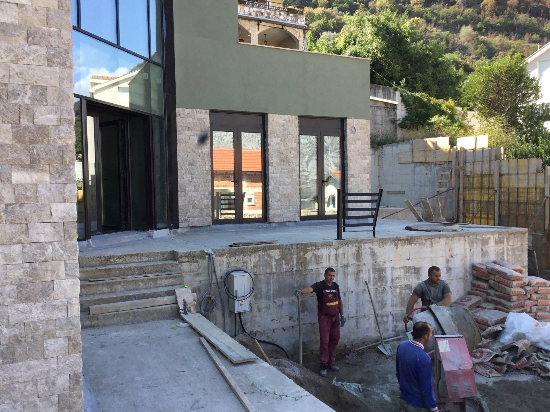Exterior of a building under construction. Two workers on a concrete platform, another pushing a wheelbarrow. Stone and glass façade.